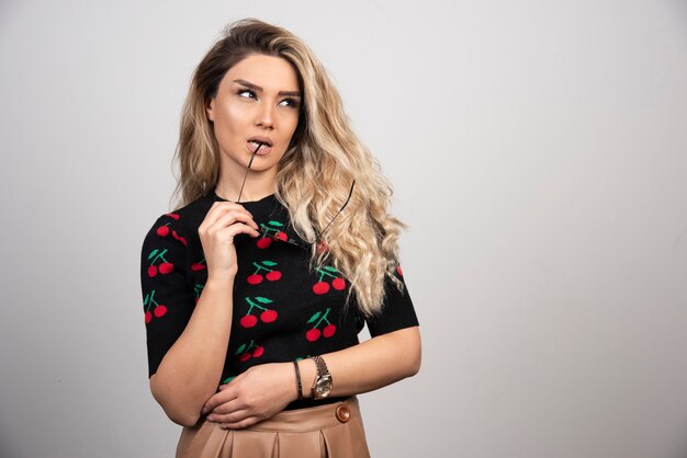 Femme blonde aux cheveux longs avec des lunettes posant sur un mur gris.