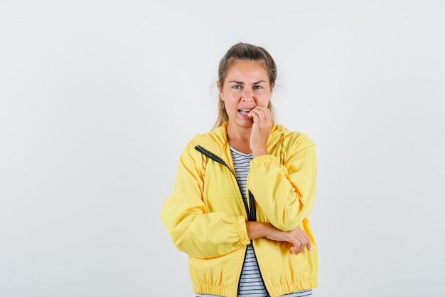 Femme blonde en blouson aviateur jaune et chemise rayée se mordant les doigts en se tenant debout dans la pose de pensée et à la pensif