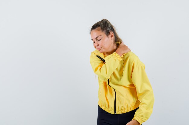 Femme blonde en blouson aviateur jaune et pantalon noir ayant des douleurs à l'épaule et à la fatigue