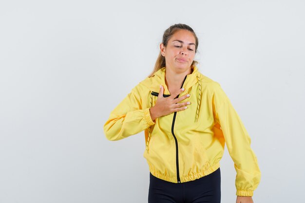 Femme blonde en blouson aviateur jaune et pantalon noir mettant la main sur la poitrine et à la sérieuse