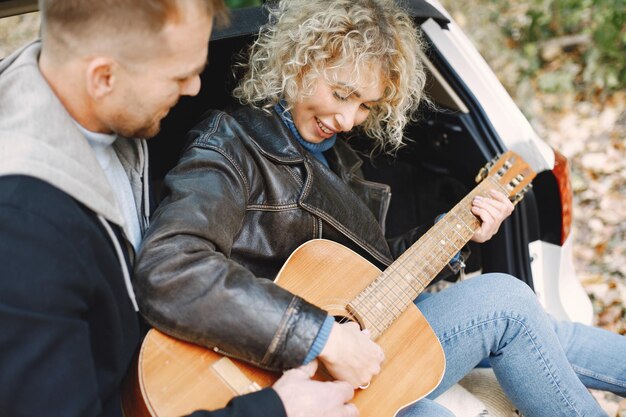 Femme blonde bouclée et homme assis dans un coffre en voiture dans la forêt d'automne avec une guitare