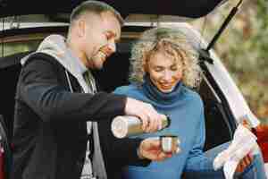 Photo gratuite femme blonde bouclée et homme assis dans un coffre en voiture dans la forêt d'automne avec une tasse thermo