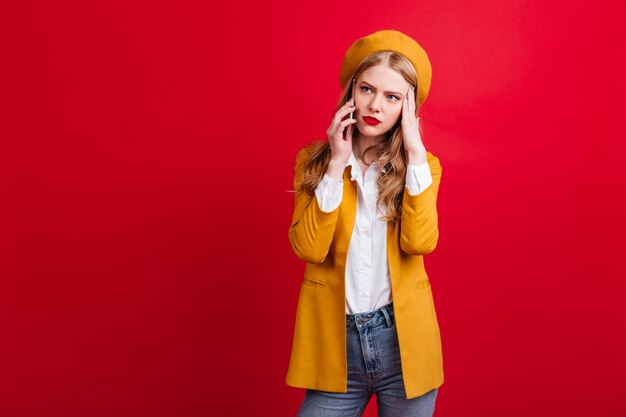 Femme blonde bouleversée en veste jaune, parler au téléphone. Triste dame caucasienne en béret debout sur un mur rouge avec smartphone.