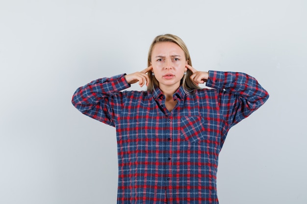 Femme blonde brancher les oreilles avec l'index en chemise à carreaux et à la colère, vue de face.