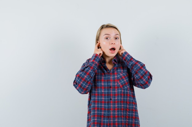 Femme blonde brancher les oreilles avec l'index en chemise à carreaux et à la surprise, vue de face.