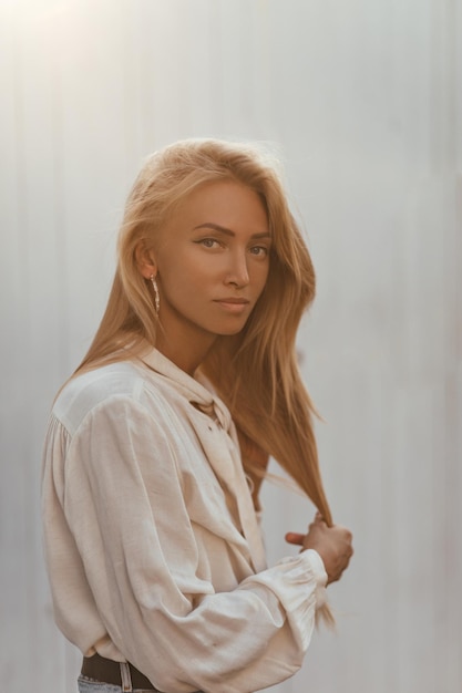 Femme blonde bronzée aux cheveux longs aux yeux bleus en chemisier en coton beige clair regarde dans la caméra et pose à l'extérieur près du mur blanc