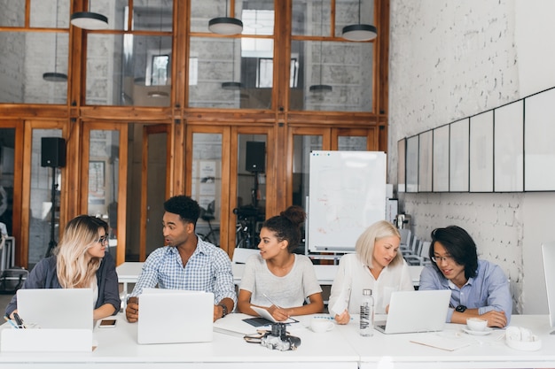 Femme blonde en chemise blanche, parler avec un ami asiatique et boire du café près d'un ordinateur portable avec tableau à feuilles mobiles. Web-designers indépendants travaillant ensemble dans une salle de conférence et utilisant des ordinateurs.