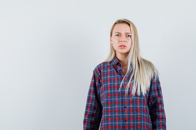 Femme blonde en chemise à carreaux debout droite et grimaçant et à la mécontentement, vue de face.