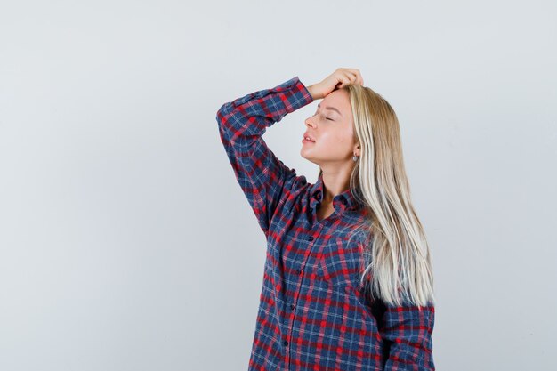 Femme blonde en chemise à carreaux mettant la main sur la tête et posant à la caméra et à la recherche attrayante, vue de face.