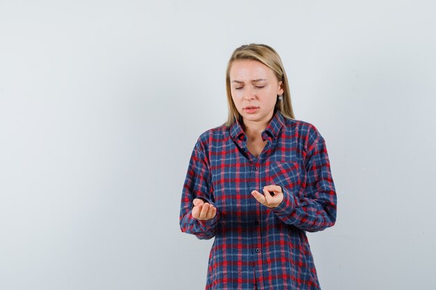 Femme blonde en chemise à carreaux regardant les mains comme tenant quelque chose et grimaçant et regardant mécontent, vue de face.