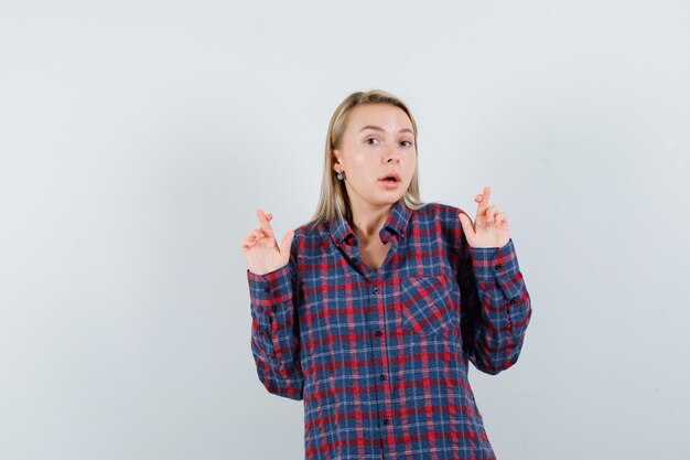 Femme blonde en chemise à carreaux tenant les doigts croisés et à la surprise, vue de face.