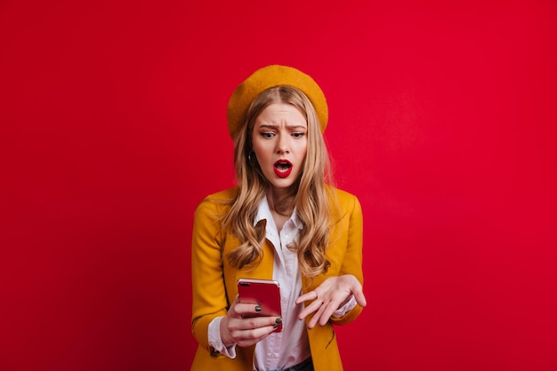 Femme blonde choquée en béret regardant smartphone Fille française bouleversée debout sur fond rouge avec la bouche ouverte