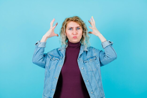 Une femme blonde en colère regarde la caméra en se tenant la main près de la tête sur fond bleu