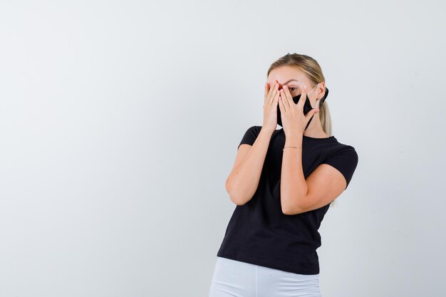 Femme blonde couvrant le visage avec les mains, regardant à travers les doigts en t-shirt noir