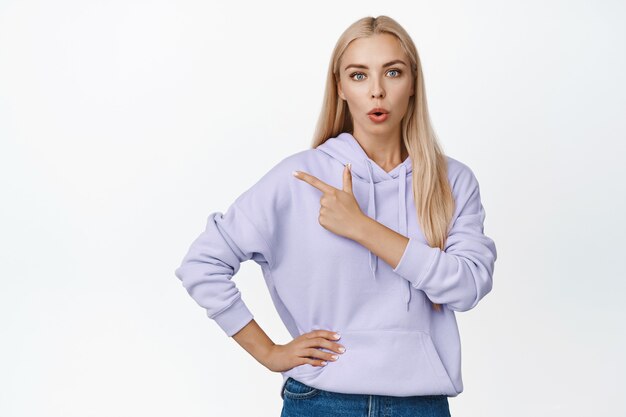 Une femme blonde curieuse pose une question sur le produit, pointant le doigt vers la gauche et l'air surpris, debout sur blanc