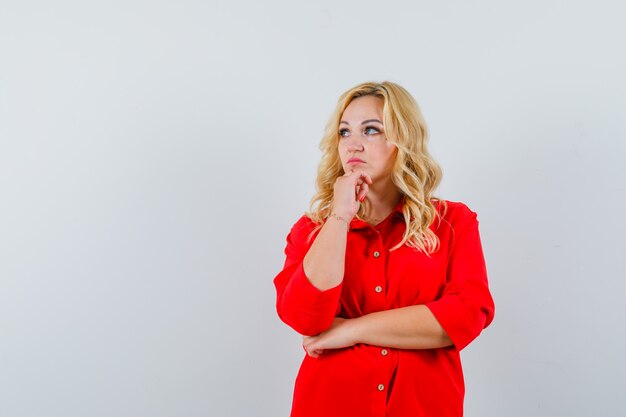 Femme blonde debout dans la pensée pose en chemisier rouge et à la pensif.