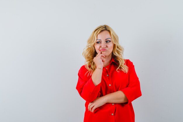 Femme blonde debout dans la pensée pose en chemisier rouge et à la pensif.