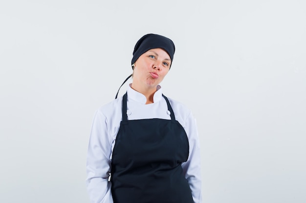 Photo gratuite femme blonde debout droite, lèvres courbes et posant à l'avant en uniforme de cuisinier noir et à la jolie.