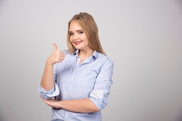 Femme blonde debout et montrant le pouce contre le mur gris.