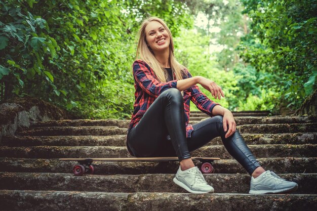 Femme blonde décontractée avec longboard utilisant un smartphone et posant dans des escaliers dans un parc naturel sauvage.