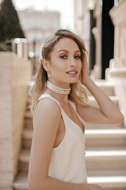 Une femme blonde élégante aux yeux gris avec un beau maquillage en collier de perles et une robe blanche se penche sur la caméra et sourit à l'extérieur