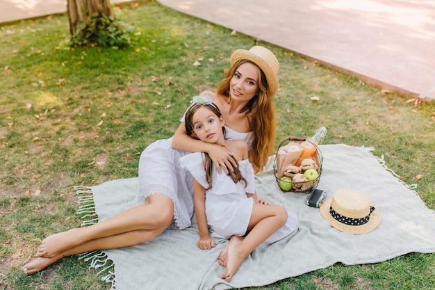 Femme blonde élégante se trouve sur une couverture blanche avec panier de pommes en week-end. Portrait en plein air de fille joyeuse et sa mère profitant du beau temps dans le parc.