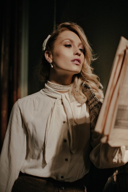 Une femme blonde élégante et sûre d'elle en blouse blanche et pantalon marron lit le journal et pose dans un couloir sombre
