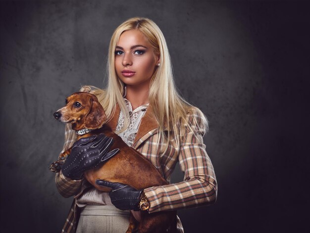 Une femme blonde élégante vêtue d'une veste à l'ancienne tient un chien blaireau rouge.