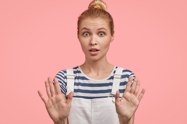 Photo gratuite une femme blonde européenne pose avec les mains tendues, démontre un geste d'arrêt, demande à s'arrêter, a surpris l'expression effrayée, exige de ne pas la déranger, montre l'interdiction, porte une salopette à la mode