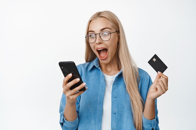 Femme blonde excitée dans des verres regardant l'écran du smartphone et criant, regardant surprise et émerveillée sur le blanc