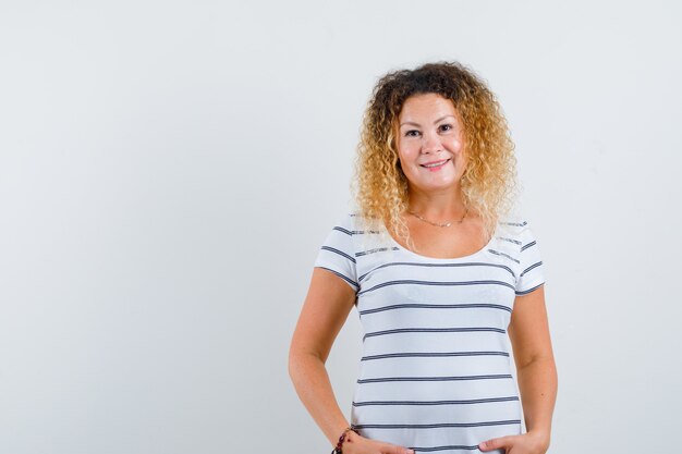 Femme blonde gardant les mains sur les hanches en t-shirt rayé et à la gaieté. vue de face.