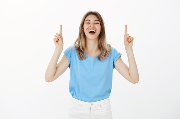 Femme blonde heureuse amusée en riant, pointant les doigts vers le haut, faisant l'annonce