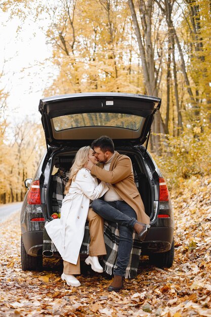 Femme blonde et homme brune assis dans un coffre de voiture noir dans la forêt d'automne. Femme portant un manteau blanc et un manteau beige pour homme. Beau couple qui s'embrasse.