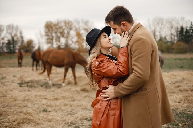 Femme blonde et homme brune debout sur le terrain avec des chevaux bruns. Femme portant une robe noire, un manteau en cuir et un chapeau et un homme portant un manteau marron. Homme et femme s'embrassant.