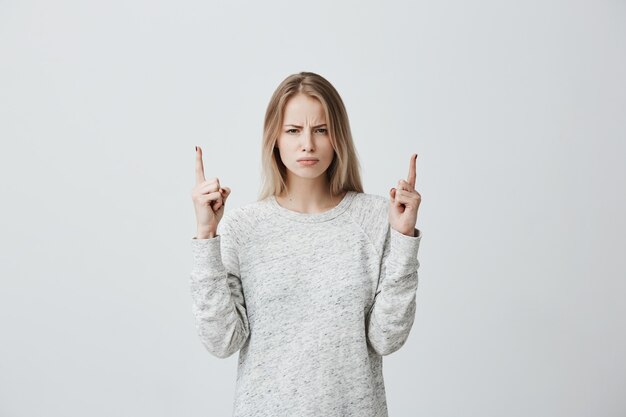 Femme blonde insatisfaite, froncer les sourcils, regardant avec colère et pointer du doigt l'espace de copie au-dessus de la tête