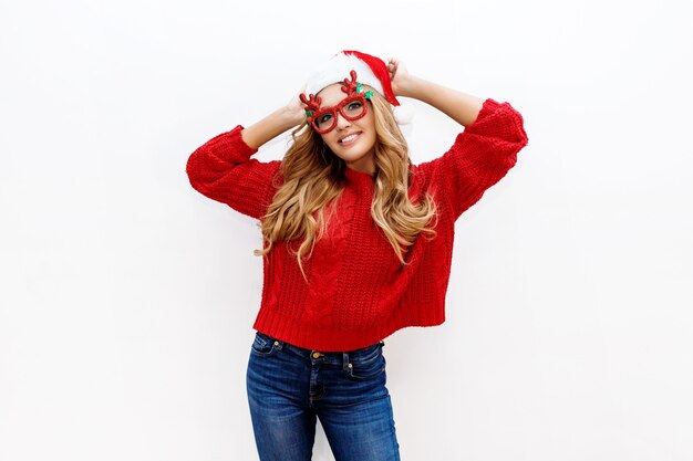 Femme blonde insouciante joyeuse dans de jolies lunettes de mascarade et chapeau de nouvel an en pull tricoté rouge posant sur un mur blanc. Isoler. Noël et nouveau concept de fête de test.