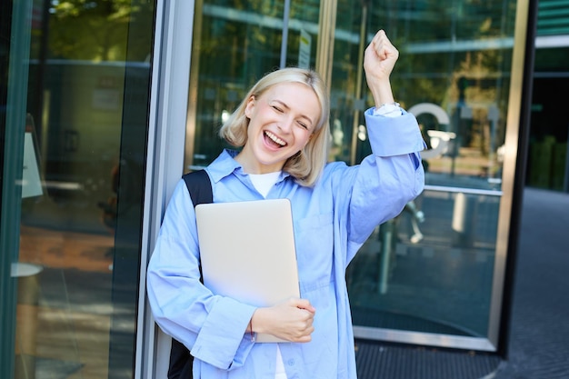 Une femme blonde insouciante qui rit, sourit et célèbre en posant avec un ordinateur portable près du bâtiment du campus du bureau