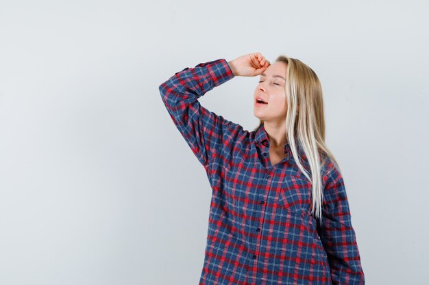 Femme blonde levant le bras et mettant la main sur la tête, posant à la caméra en chemise à carreaux et à la recherche attrayante. vue de face.