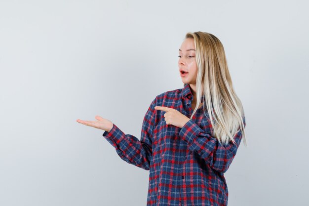Femme blonde levant les mains comme tenant quelque chose et pointant vers elle avec une autre main en chemise à carreaux et à la vue de face, focalisée