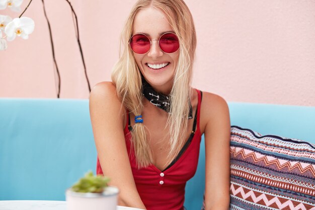 femme blonde en lunettes de soleil à la mode, porte des vêtements à la mode et des lunettes de soleil rouges, s'assoit contre le mur rose sur un canapé confortable.