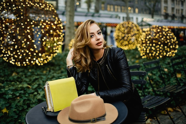 Une Femme Blonde Magnifique Se Trouve à La Table Ronde Noire Avant Les Buissons Verts à L'extérieur