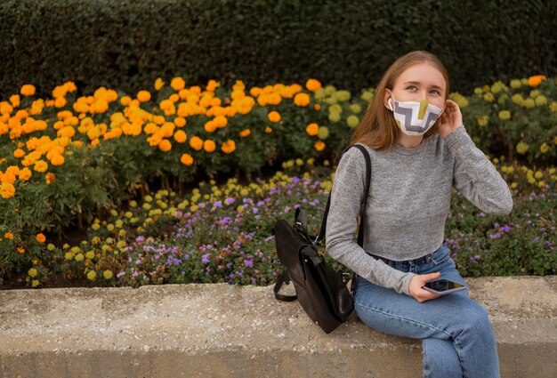 Femme blonde avec masque médical assis à côté d'un jardin avec espace copie