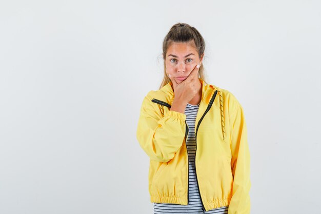 Femme blonde mettant la main sur le menton tout en pensant poser en blouson aviateur jaune et chemise rayée et à la pensif