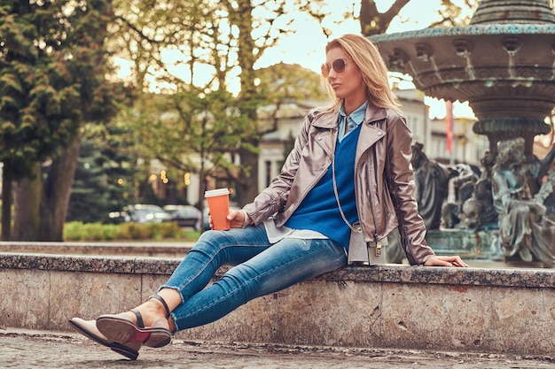 Une femme blonde à la mode se détend en plein air, buvant du café à emporter tout en étant assise sur le banc du parc de la ville.