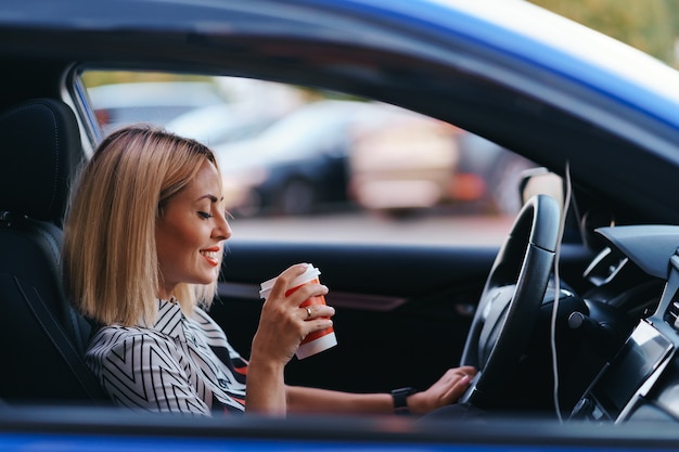 Femme blonde moderne ayant un café à emporter en conduisant dans la ville