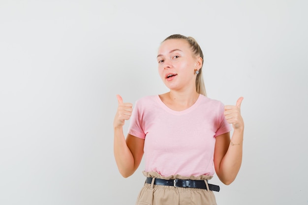 Femme blonde montrant deux pouces vers le haut en t-shirt, pantalon et regardant jovial, vue de face.