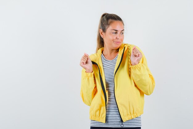 Femme blonde montrant le geste du gagnant en blouson aviateur jaune et chemise rayée et à la recherche de plaisir