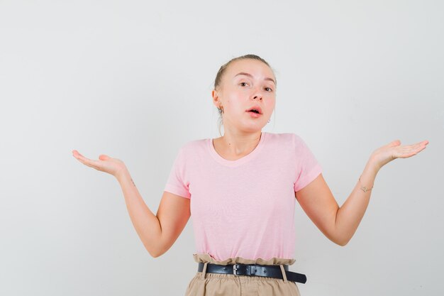 Femme blonde montrant un geste impuissant en t-shirt, pantalon et à la confusion, vue de face.