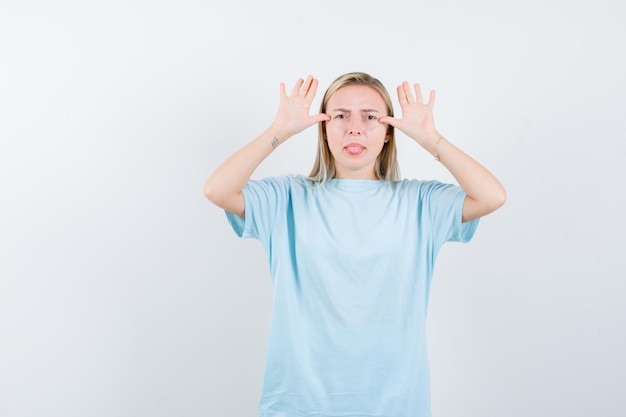 Femme blonde montrant le geste des oreilles, tirant la langue en t-shirt bleu et l'air amusé