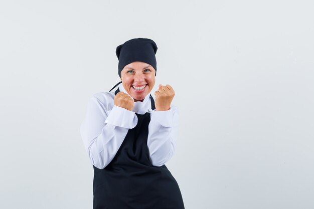 Femme blonde montrant le geste de réussite en uniforme de cuisinier noir et à la recherche de plaisir. vue de face.